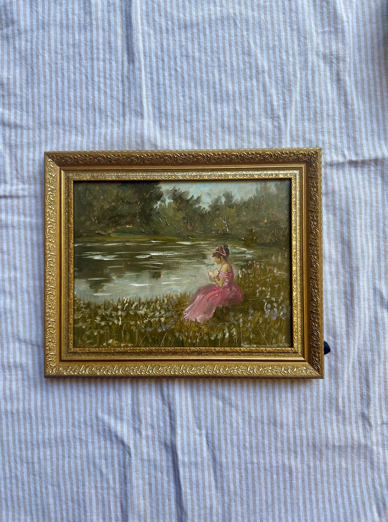 Framed painting of a woman in a pink dress by a lake on a textured fabric background