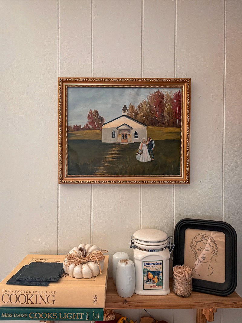 Framed painting of a small building on a wall above a wooden shelf with decorative items.